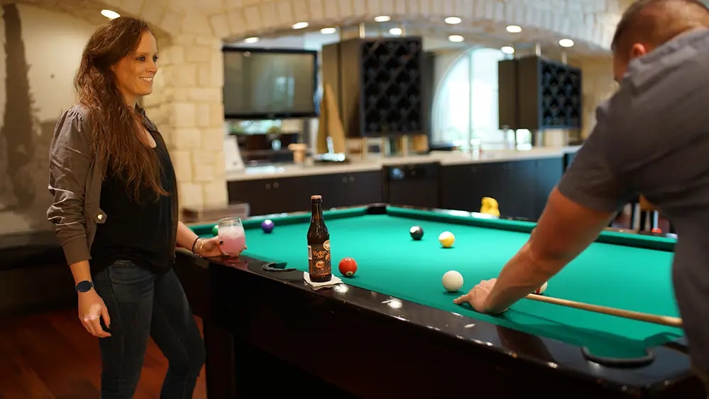 A lady watching a gentleman play pool, A beer is on the table with a Panic Button on her wrist.