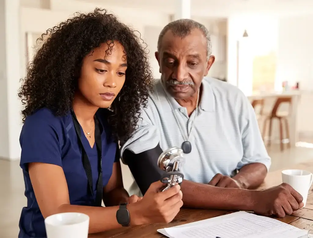 Healthcare professional assisting an older man with a blood pressure cuff, both seated at a table. The healthcare professional is wearing a Silent Beacon device.